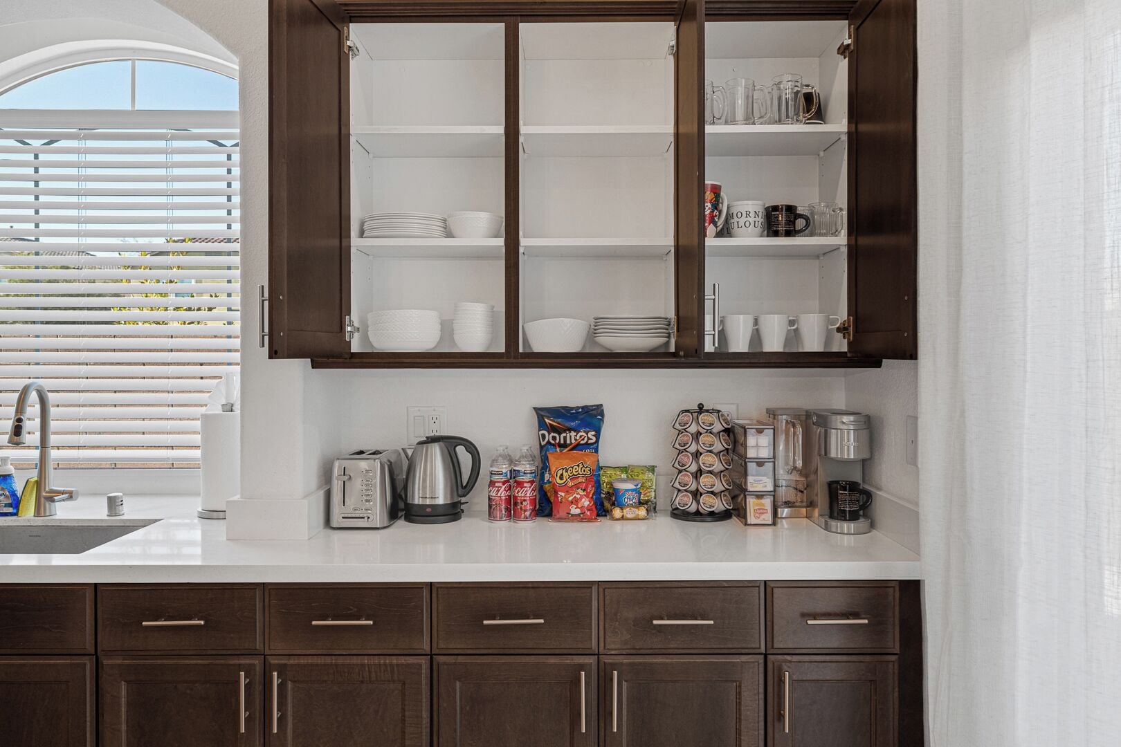 Una cocina con encimera blanca, armarios de madera oscura y una ventana con persianas.