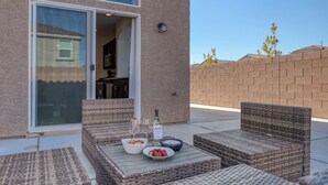 Een terras buiten met rieten meubels, een tafel met een fles en glazen, en een schaal fruit.