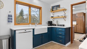 Een moderne keuken met een vaatwasser, spoelbak en blauwe kastjes. Daarboven staan houten planken bij de spoelbak en een schoolbord met een tekening van een schip.