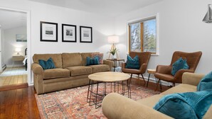 Een woonkamer met een bruine bank, twee fauteuils, een salontafel en een tapijt met een patroon. Er hangen ingelijste foto's aan de muur, een lamp en een raam met uitzicht op bomen.