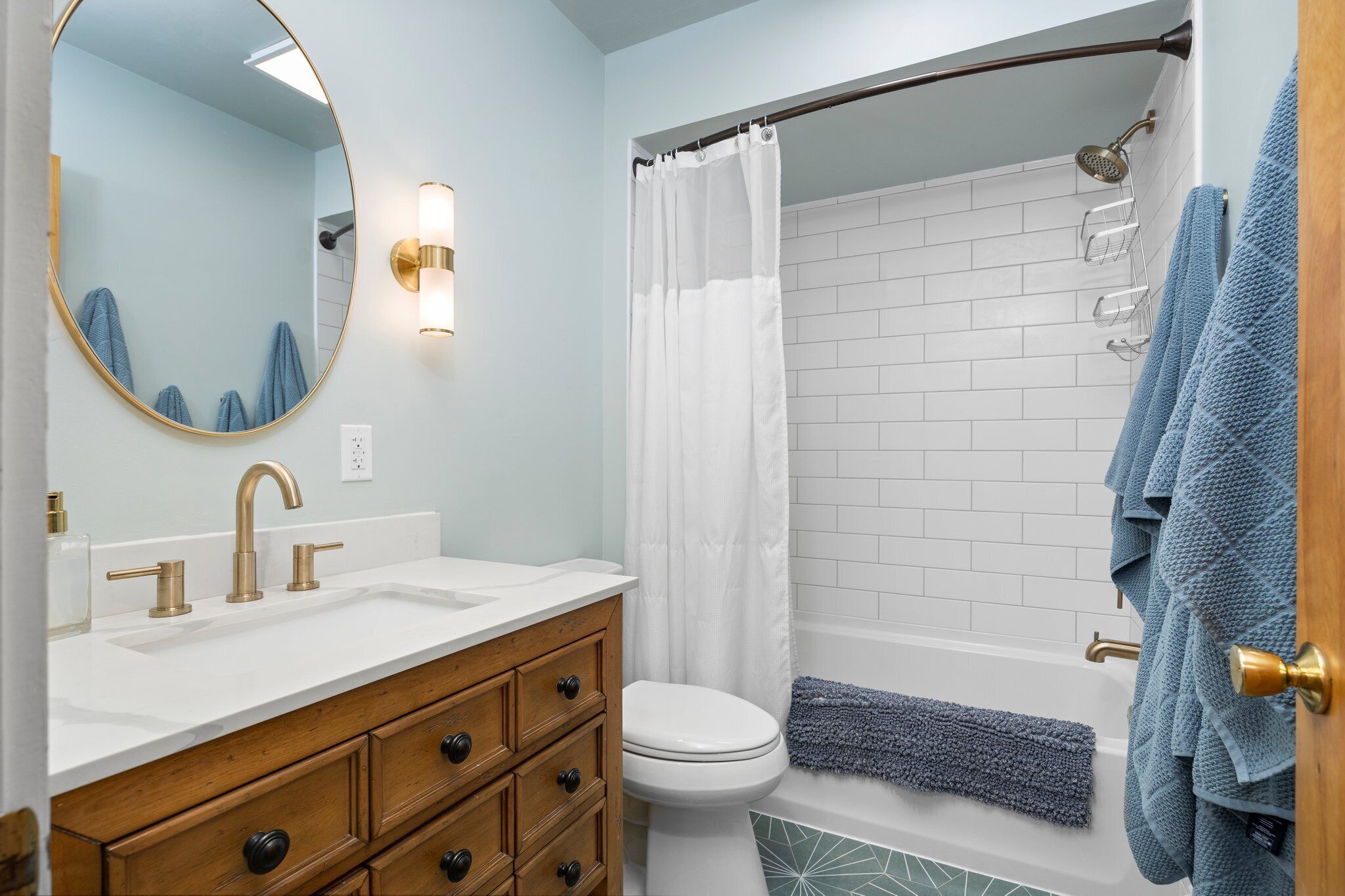A bathroom with a white sink, gold faucets, a wooden vanity, a white toilet, a shower with a white curtain, and blue towels.