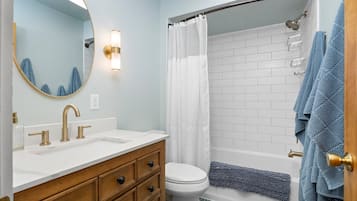 A bathroom with a white sink, gold faucets, a wooden vanity, a white toilet, a shower with a white curtain, and blue towels.