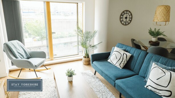 A modern living room with a blue sofa, a rocking chair, and a large window with a view of the city.