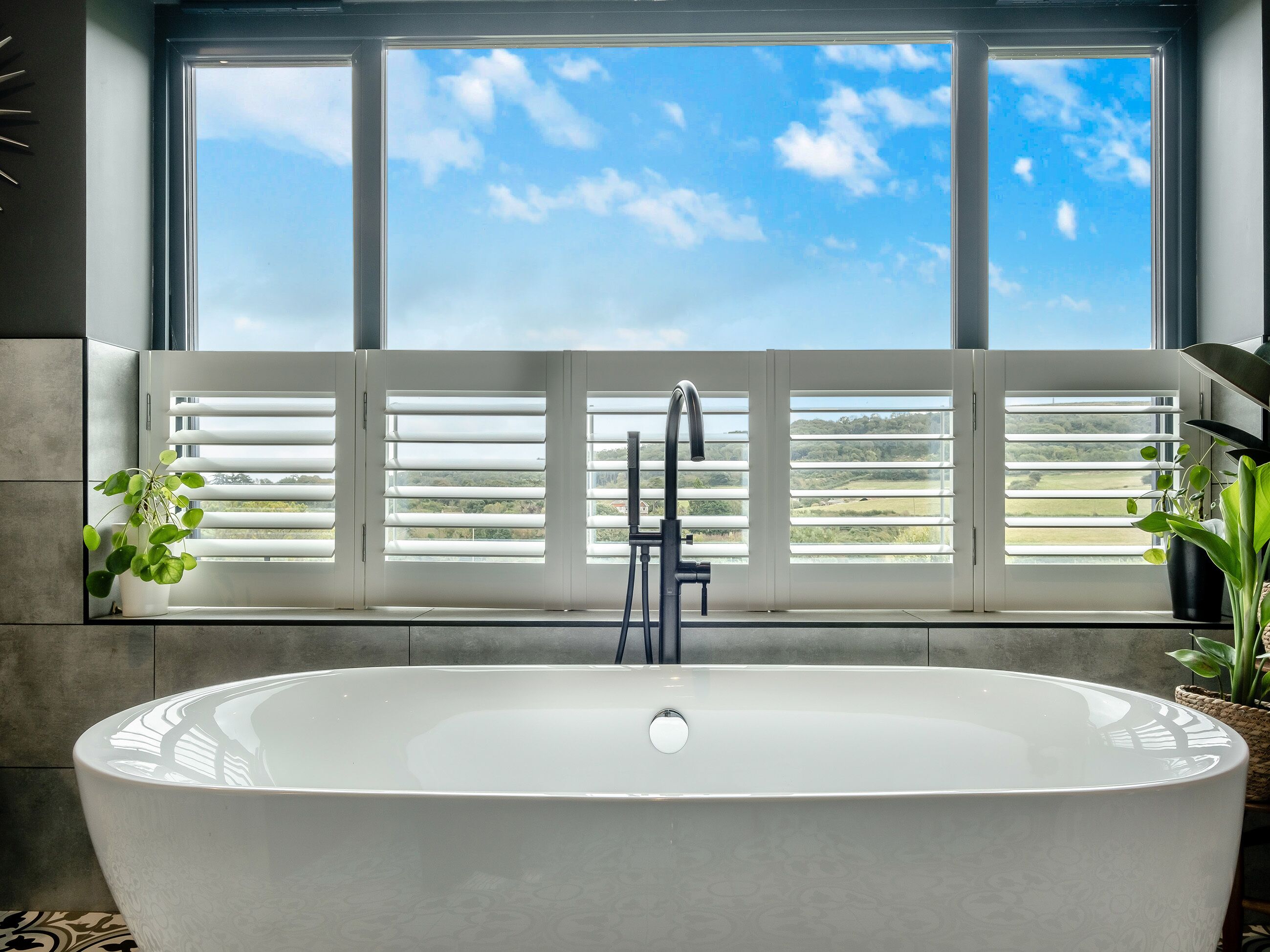 A modern bathroom with a freestanding bathtub, a window with blinds, and potted plants.