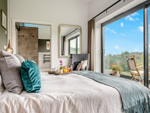 A modern hotel room with a large bed, a dining area with breakfast, a large mirror, and a balcony with a view.
