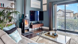 A modern living room with a fireplace, a flat-screen TV, and a large window offering a view of a house under construction. - 5 bedroom accommodation in Totland (Totland)