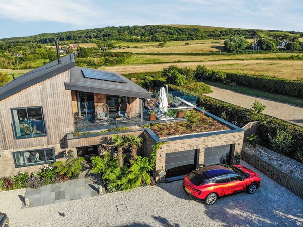 A modern house with a flat roof, a covered outdoor seating area, and a red sports car parked in the driveway.