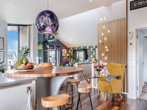 A modern kitchen with a central island, wooden bar stools, a yellow armchair, and a decorative hanging light fixture.