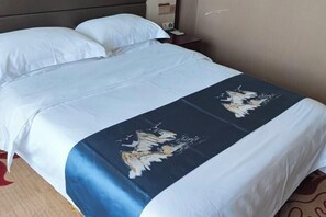 Quarto de hotel com uma cama, roupa de cama branca, colcha abstrata azul e branca e mesa de cabeceira com abajur.