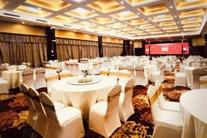 Une salle de banquet avec des tables rondes, des chaises blanches et une scène surmontée d’un rideau rouge.
