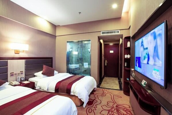 Quarto de hotel com duas camas, televisão, minibar e banheiro visível através de uma porta de vidro.