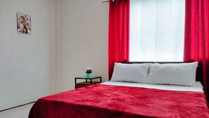 A bedroom with a bed, red curtains, a small table with a plant, and a wall-mounted picture. - Reddoorz @ Rio Bella Lacson St. Bacolod (Bacolod)