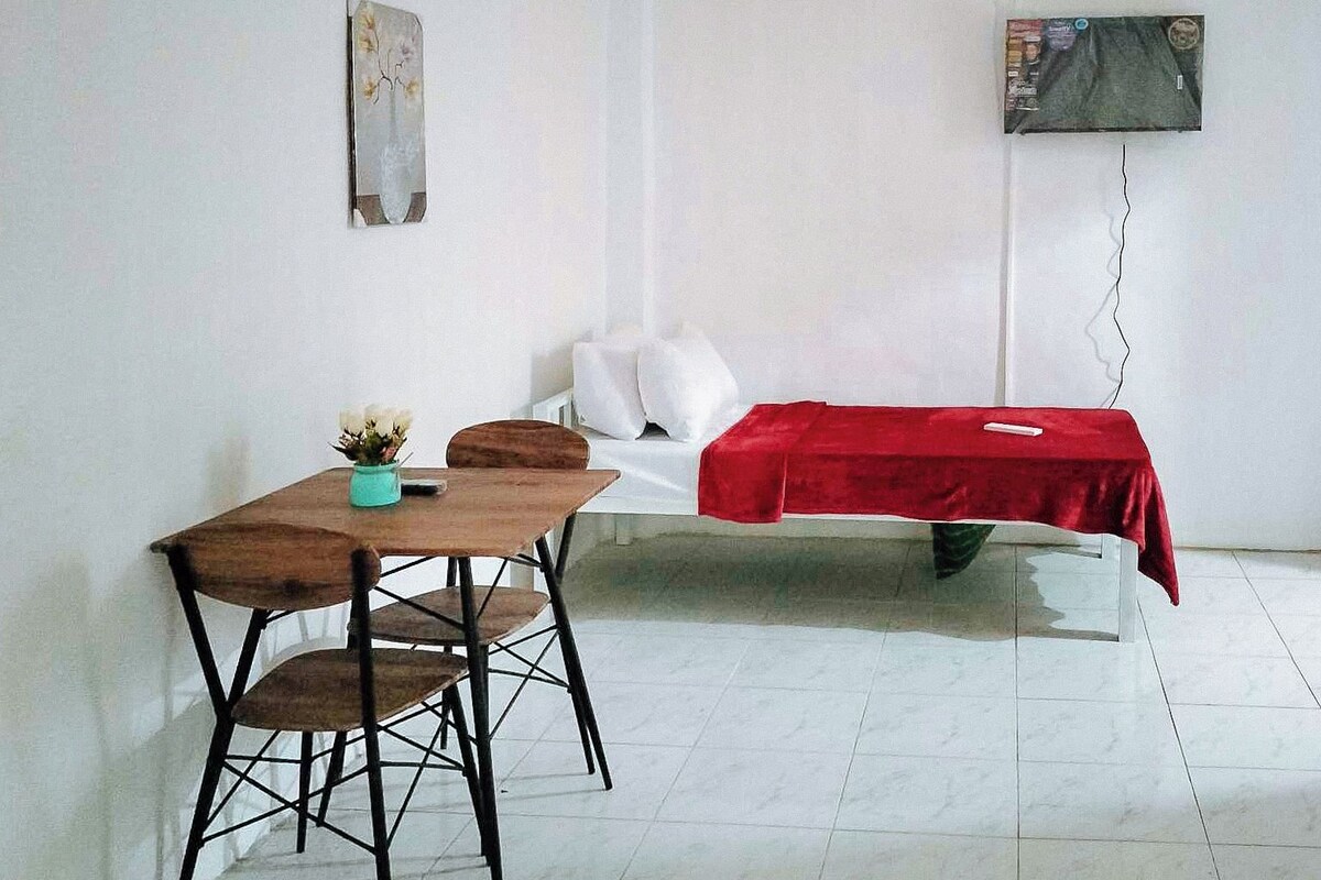 A room with a bed covered in a red blanket, a small table with a chair, and a wall-mounted TV.