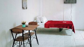 A room with a bed covered in a red blanket, a small table with a chair, and a wall-mounted TV.