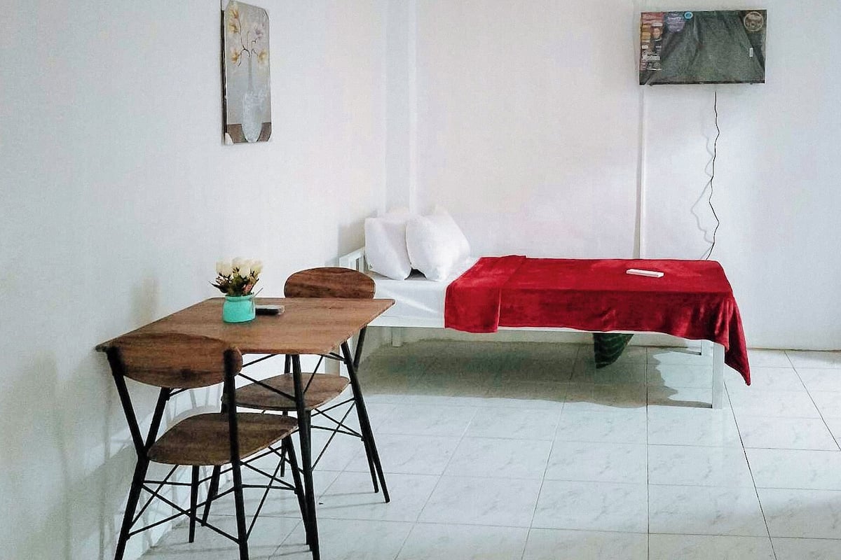Una habitación con una cama cubierta por una colcha roja, una mesita con una silla y un televisor montado en la pared.