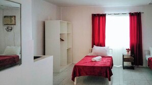 Une chambre d’hôtel avec un lit, une armoire, une petite table sur laquelle se trouve un vase, et une fenêtre avec des rideaux rouges.