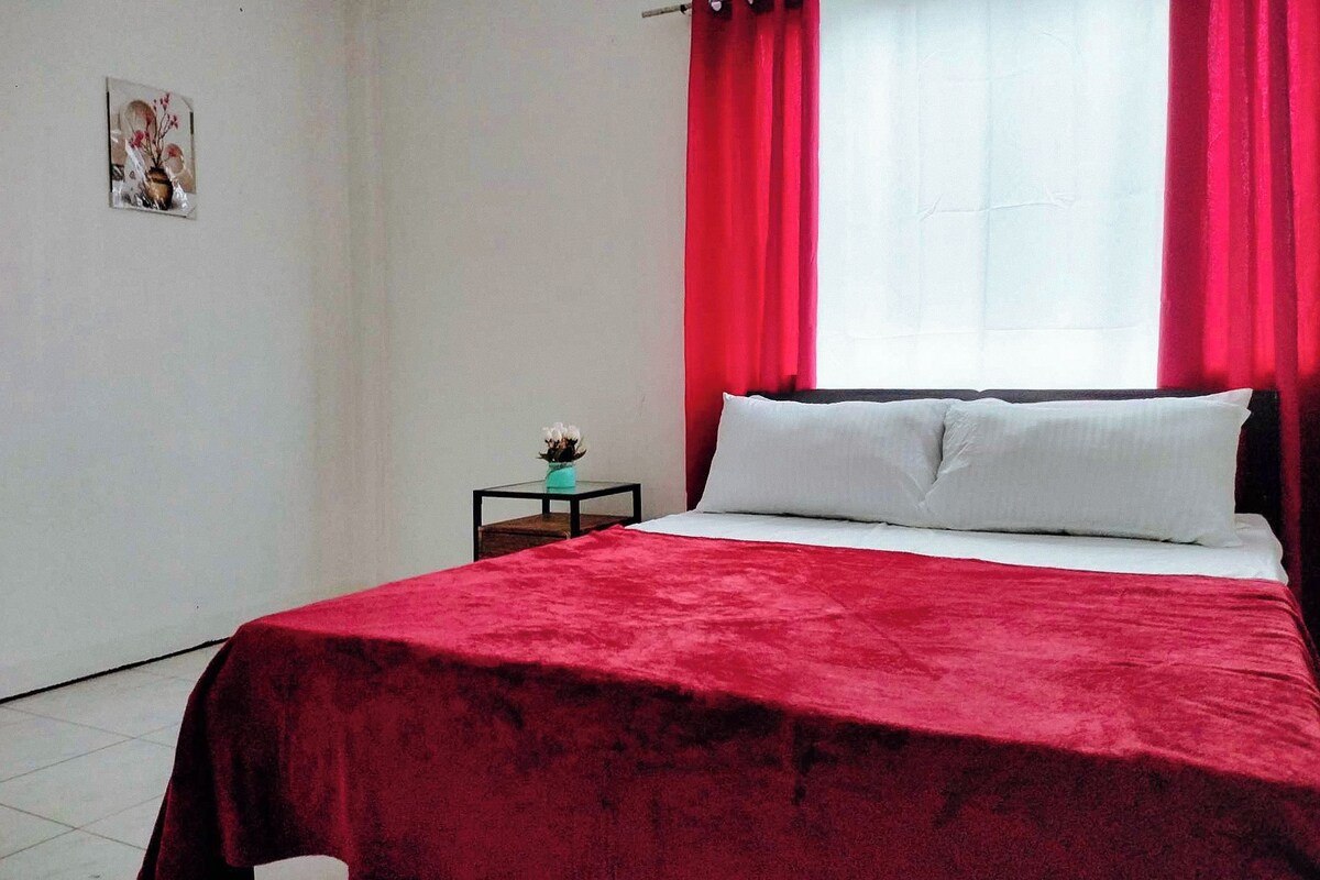A bedroom with a bed, red curtains, a small table with a plant, and a wall-mounted picture.
