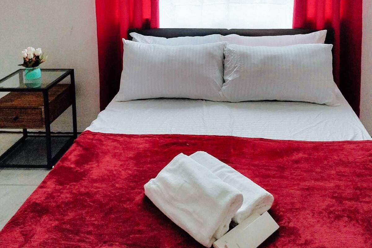 Una cama bien hecha con sábanas y almohadones blancos, una manta roja y una toalla blanca doblada sobre la cama.