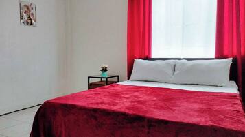 A bedroom with a bed, red curtains, a small table with a plant, and a wall-mounted picture.