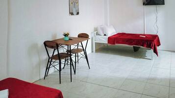 A room with a dining table, two chairs, a bed with a red blanket, a white sofa, and a wall-mounted TV.