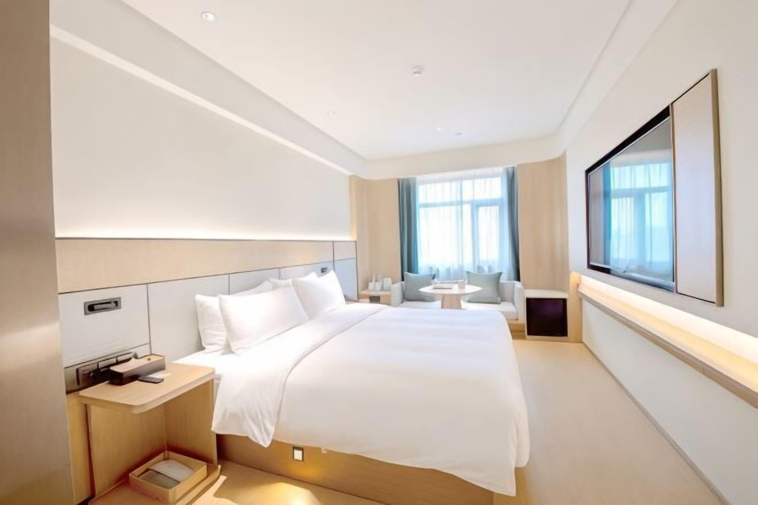 A modern hotel room with a large bed, bedside tables, a TV, and a window with curtains.