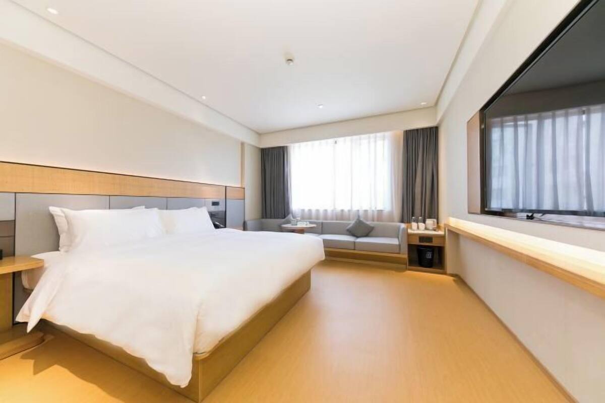 A modern hotel room with a large bed, a sofa, a small table, and a window with curtains.