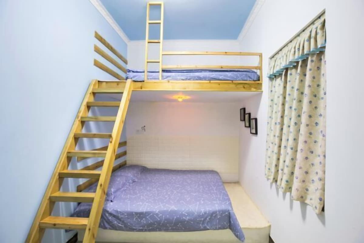 A bunk bed room with a wooden ladder, a window with curtains, and a wall-mounted light fixture.