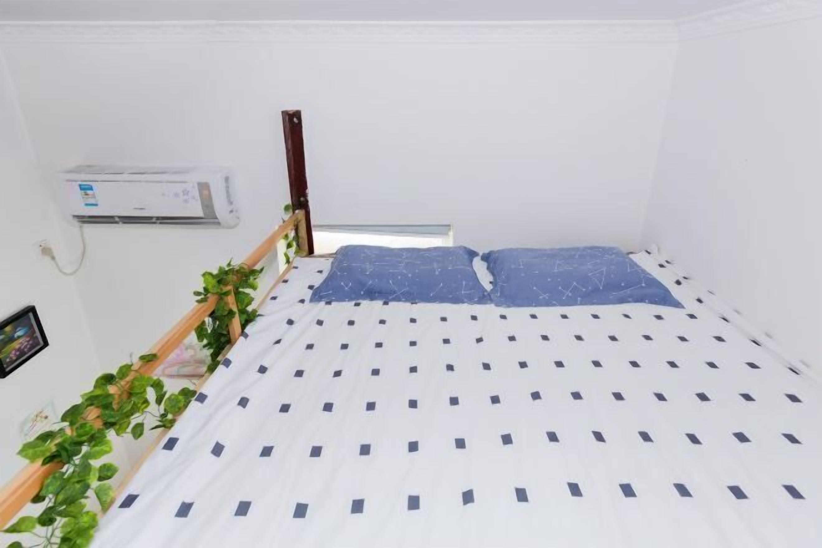 Una habitación pequeña con cama individual, cubrecolcha blanca y azul estampada, cabecera de madera, unidad de aire acondicionado en la pared y una planta en maceta a la izquierda.