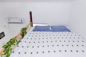 A small, single-bed room with a white and blue patterned bedspread, a wooden headboard, an air conditioning unit on the wall, and a potted plant on the left side.