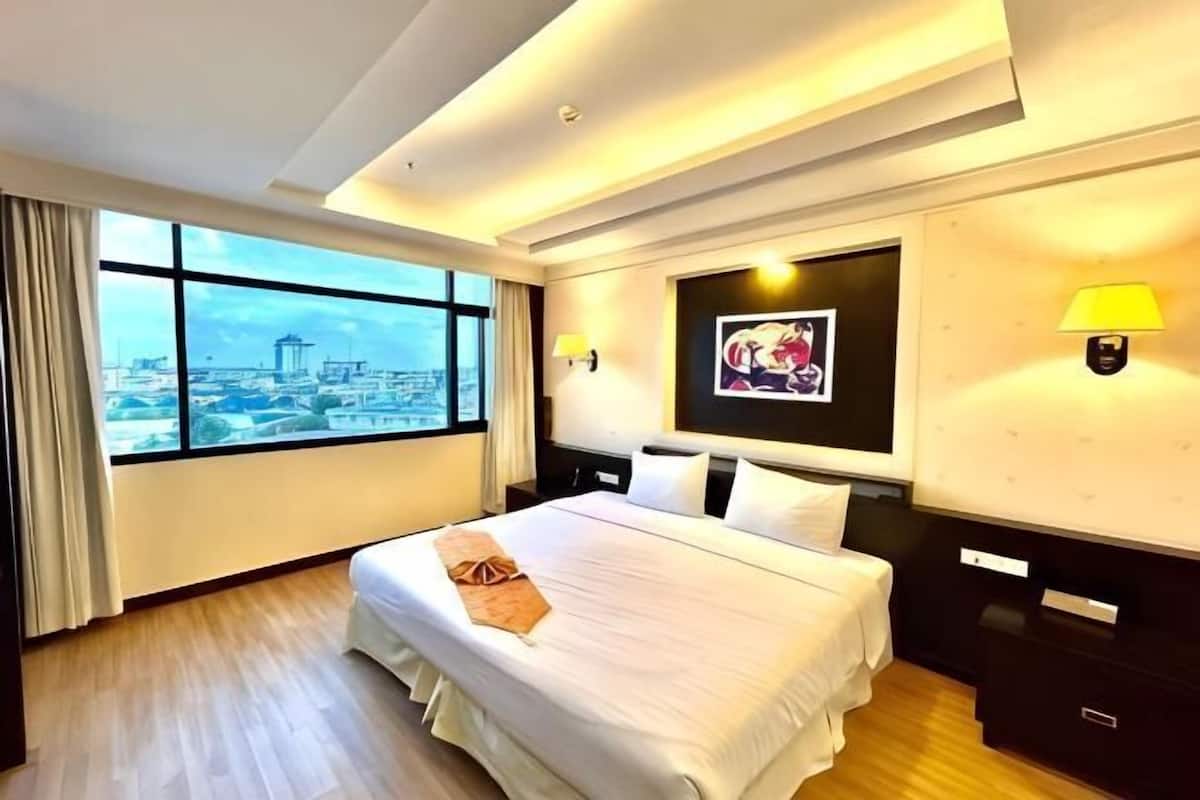 A hotel room with a large bed, a city view, a framed picture on the wall, and a nightstand with a lamp.