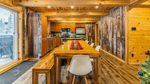 A wooden interior with a long dining table, chairs, and a kitchen area. - The Escapade - Private hot tub with pool table 6BR (Sainte-Lucie-des-Laurentides)