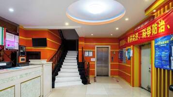 A hotel lobby with a reception desk, a staircase, and a red wall with informational posters.