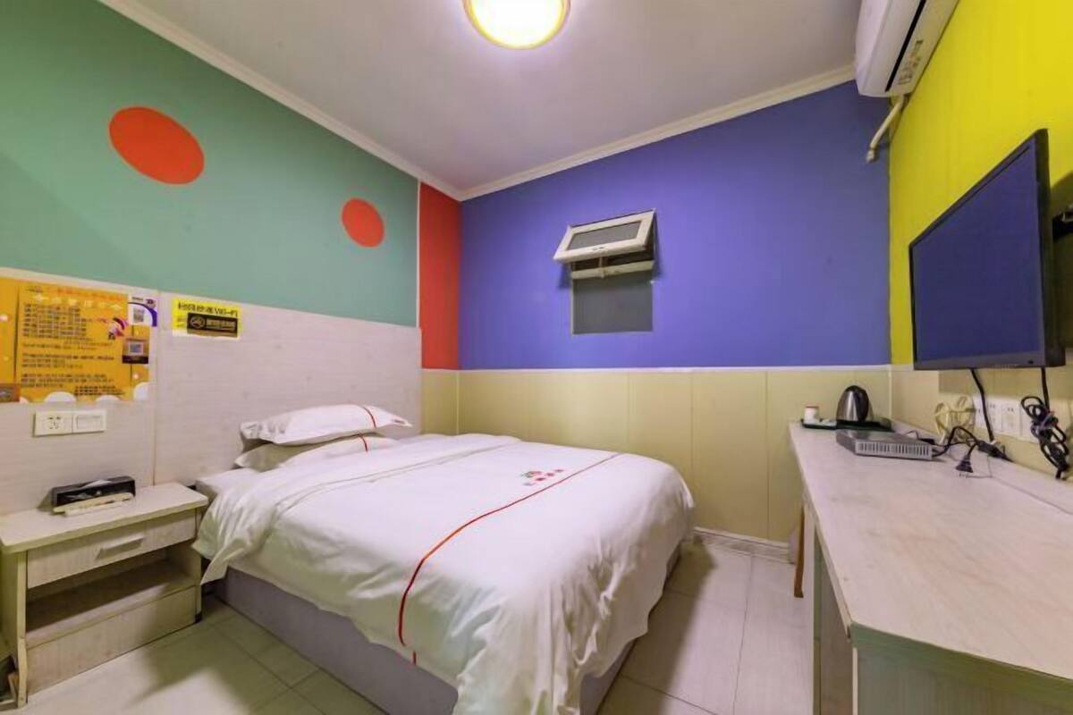 A room with a bed, a bedside table, a small desk, an air conditioning unit, a mirror, and a wall with colorful circles.