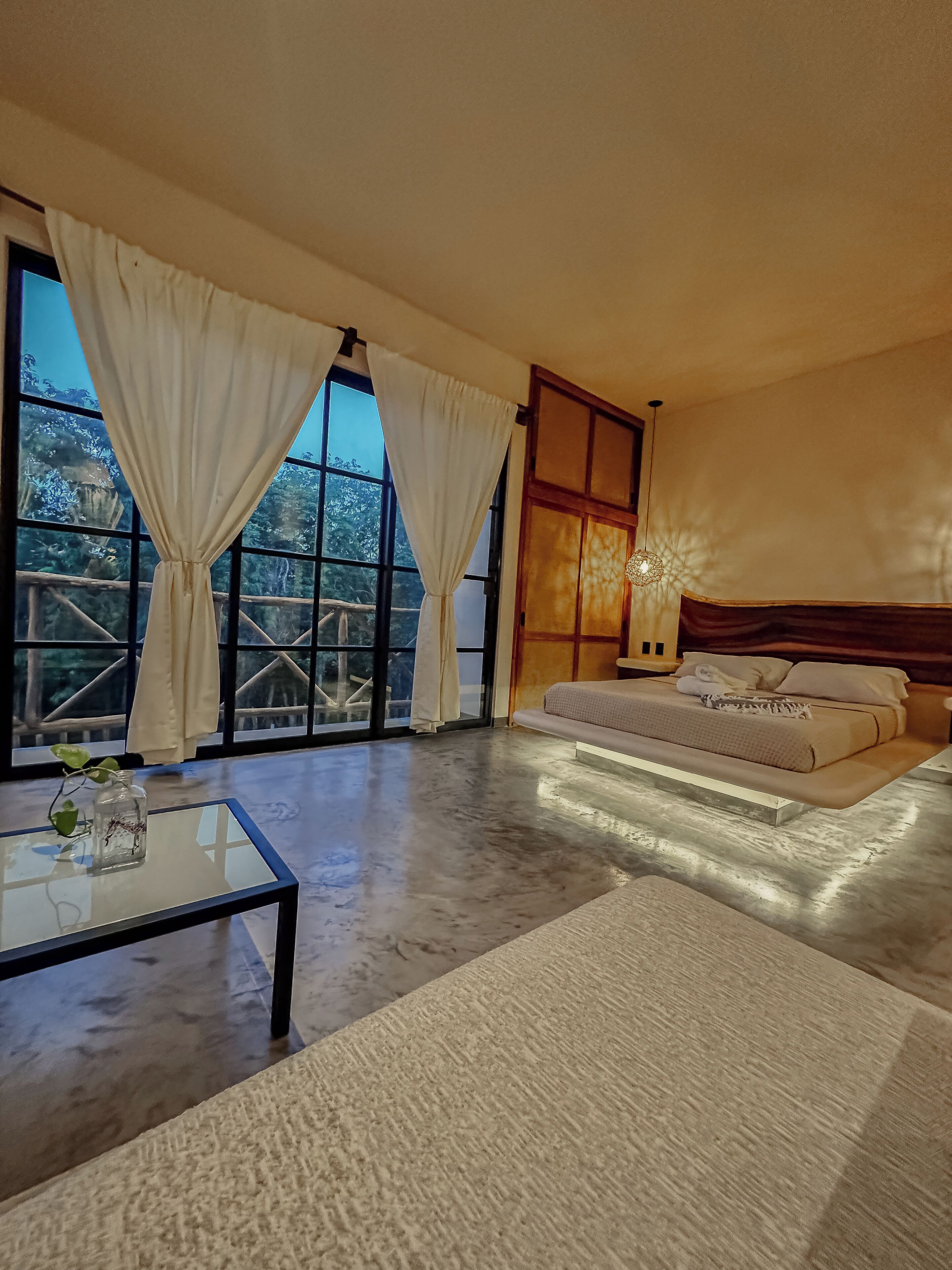 A modern bedroom with a large bed, a glass coffee table, and a view of a forest through large windows.