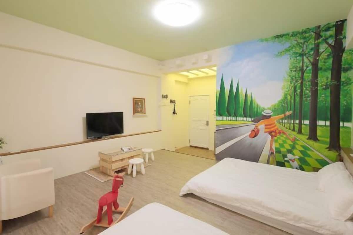 A hotel room with a television, a desk, a chair, a bed, and a mural of a forest scene.
