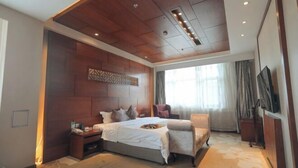Habitación de hotel moderna con techo de madera, una cama grande con almohadas, una silla, un televisor y una ventana con cortinas.