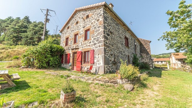 A stone building with a red door and windows, surrounded by a grassy area and trees.