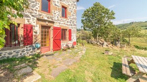 A stone house with red shutters and a stone pathway leading to a wooden bench.