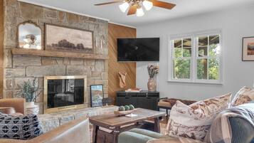 A living room with a stone fireplace, a television mounted on the wall, and a large window with a view of trees.