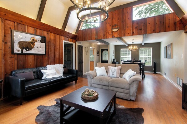 A spacious living area with wooden walls, a black leather sofa, a grey sofa, and a dining table with chairs. - Hidden Cabin Retreat with Private Deck & Firepit (Gainesville)
