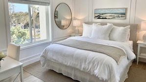 A bedroom with a large bed, a round mirror, a painting, and a view of trees and a street through the window.