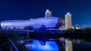 A modern building with a unique curved design, reflecting in a calm body of water, surrounded by greenery and other buildings in the background. - Yijing International Hotel (Fuyang)
