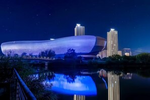 A modern building with a unique curved design, reflecting in a calm body of water, surrounded by greenery and other buildings in the background. - Yijing International Hotel (Fuyang)