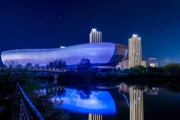 A modern building with a unique curved design, reflecting in a calm body of water, surrounded by greenery and other buildings in the background. - YIJING International Hotel (Fuyang)