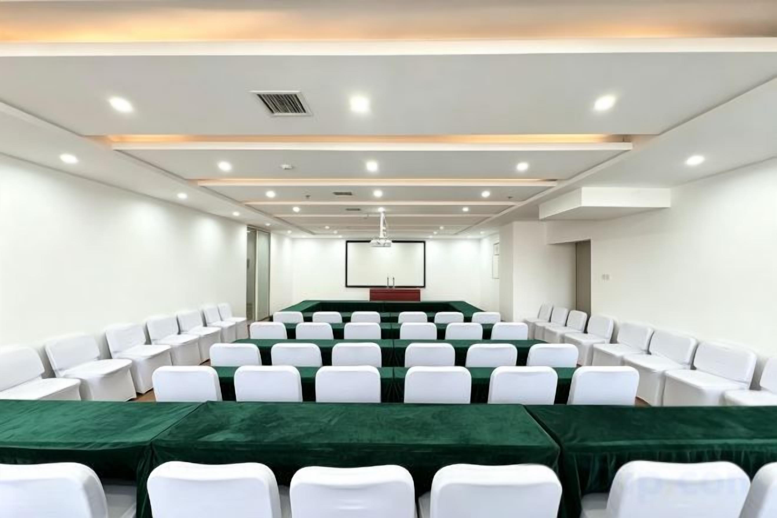 A conference room with rows of white chairs, a long table with a green tablecloth, and a projector screen at the front.
