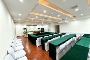 Un salón de conferencias con largas mesas y sillas blancas, una pantalla de proyector y una planta.