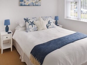 Cama cuidadosamente hecha con almohadas de estrella de mar, una manta azul y cabecera blanca. Dos mesitas de noche, una con lámpara y un teléfono inteligente. Un cuadro en la pared sobre la cama.