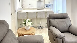 Un salon moderne avec deux canapés gris, une petite table ronde en bois surmontée d’un vase blanc, et une cuisine en arrière-plan.