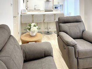 Ein modernes Wohnzimmer mit zwei grauen Sofas, einem kleinen runden Holztisch mit einer weißen Vase und einer Küche im Hintergrund.