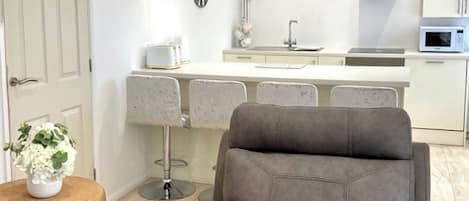 Cocina moderna con armarios blancos, microondas y campana extractora de acero inoxidable. Hay un comedor con una silla tapizada de color gris y una mesa de mimbre con una planta en maceta.